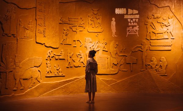 Visiteuse curieuse devant une murale de hiéroglyphes illustrant les questions qui révèlent notre monde 